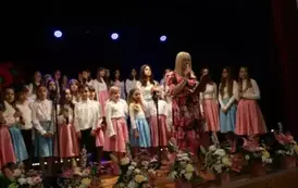 Grupa dorosłych os&oacute;b stoi razem na scenie, pozując do pamiątkowego zdjęcia. Większość z nich trzyma w rękach r&oacute;że. W tle czarna kurtyna i niebieskie światła sceniczne. Na pierwszym planie widoczne są kompozycje kwiatowe. Atmosfera wskazuje na oficjalne wy