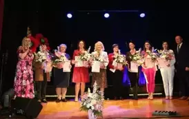 Grupa dorosłych os&oacute;b stoi razem na scenie, pozując do pamiątkowego zdjęcia. Większość z nich trzyma w rękach r&oacute;że. W tle czarna kurtyna i niebieskie światła sceniczne. Na pierwszym planie widoczne są kompozycje kwiatowe. Atmosfera wskazuje na oficjalne wy
