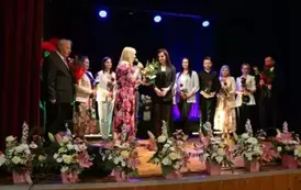 Grupa dorosłych os&oacute;b stoi razem na scenie, pozując do pamiątkowego zdjęcia. Większość z nich trzyma w rękach r&oacute;że. W tle czarna kurtyna i niebieskie światła sceniczne. Na pierwszym planie widoczne są kompozycje kwiatowe. Atmosfera wskazuje na oficjalne wy