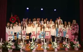 Na scenie występuje grupa dzieci i młodzieży ubranych w białe koszule i kolorowe sp&oacute;dnice (r&oacute;żowe i niebieskie). Śpiewają wsp&oacute;lnie, stojąc w kilku rzędach przed mikrofonami. Scena ozdobiona jest kompozycjami kwiatowymi w r&oacute;żowych doniczkach. Tło jest ciem