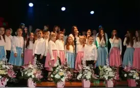 Na scenie występuje grupa dzieci i młodzieży ubranych w białe koszule i kolorowe sp&oacute;dnice (r&oacute;żowe i niebieskie). Śpiewają wsp&oacute;lnie, stojąc w kilku rzędach przed mikrofonami. Scena ozdobiona jest kompozycjami kwiatowymi w r&oacute;żowych doniczkach. Tło jest ciem