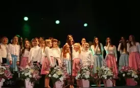 Na scenie występuje grupa dzieci i młodzieży ubranych w białe koszule i kolorowe sp&oacute;dnice (r&oacute;żowe i niebieskie). Śpiewają wsp&oacute;lnie, stojąc w kilku rzędach przed mikrofonami. Scena ozdobiona jest kompozycjami kwiatowymi w r&oacute;żowych doniczkach. Tło jest ciem
