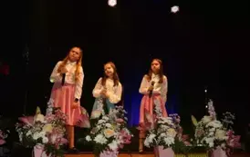 Na scenie występuje grupa dzieci i młodzieży ubranych w białe koszule i kolorowe sp&oacute;dnice (r&oacute;żowe i niebieskie). Śpiewają wsp&oacute;lnie, stojąc w kilku rzędach przed mikrofonami. Scena ozdobiona jest kompozycjami kwiatowymi w r&oacute;żowych doniczkach. Tło jest ciem