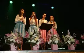 Na scenie występuje grupa dzieci i młodzieży ubranych w białe koszule i kolorowe sp&oacute;dnice (r&oacute;żowe i niebieskie). Śpiewają wsp&oacute;lnie, stojąc w kilku rzędach przed mikrofonami. Scena ozdobiona jest kompozycjami kwiatowymi w r&oacute;żowych doniczkach. Tło jest ciem