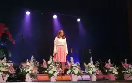 Dziewczynka spiewajaca na scenie pełnej kwiat&oacute;w. jest ubrana w Biała bluzkę i r&oacute;żową sp&oacute;dnice. 