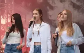 Sześć młodych os&oacute;b &ndash; pięć dziewczyn i jeden chłopak &ndash; stoi na scenie, trzymając mikrofony. Mają na sobie białe koszule lub bluzki i niebieskie jeansy. W tle widoczny sprzęt muzyczny i kolorowe światła sceniczne.