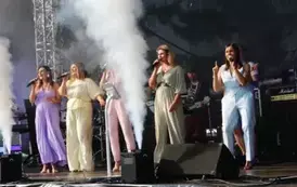 Pięć kobiet w pastelowych kombinezonach śpiewa na scenie, trzymając mikrofony. Za nimi widać muzyk&oacute;w grających na instrumentach. Oświetlenie sceniczne i sprzęt muzyczny tworzą tło.