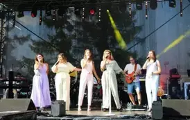 Pięć kobiet w pastelowych kombinezonach śpiewa na scenie, trzymając mikrofony. Za nimi widać muzyk&oacute;w grających na instrumentach. Oświetlenie sceniczne i sprzęt muzyczny tworzą tło.