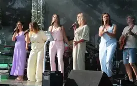 Pięć kobiet w pastelowych kombinezonach śpiewa na scenie, trzymając mikrofony. Za nimi widać muzyk&oacute;w grających na instrumentach. Oświetlenie sceniczne i sprzęt muzyczny tworzą tło.