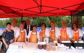 Grupa os&oacute;b stojąca pod czerwonym namiotem, za stołem z jedzeniem i napojami. Kilka kobiet nosi czarne fartuchy z napisem &bdquo;KGW i nazwa miejscowości&rdquo;, w środku mężczyzna w garniturze. W tle zielone drzewa.