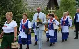 Poch&oacute;d dożynkowy. Korow&oacute;d wielu os&oacute;b ubranych elegancko, a także w stroje ludowe. Kobiety niosą dożynkowe wieńce. Niosą także chleby