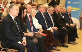 Widok na pierwszy rząd widowni w sali widowiskowej. Siedzą w nim zaproszeni goście, w tym mężczyźni w garniturach oraz kobieta w bordowej, koronkowej sukience. Po lewej stronie widać fragment postaci w stroju ludowym. W tle wypełniona sala.
