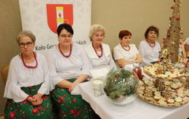 Kobiety w strojach stylizowanych na ludowe siedzą za białym stołem biesiadnym. Na stole widać dzbanek soku, p&oacute;łmiski z jedzeniem oraz sztućce. W tle banery z herbem oraz napisem &bdquo;Gmina Chork&oacute;wka&rdquo; i &bdquo;Koło Gospodyń Wiejskich&rdquo;.