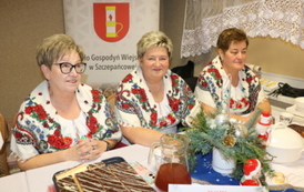 Kobiety w strojach stylizowanych na ludowe siedzą za białym stołem biesiadnym. Na stole widać dzbanek soku, p&oacute;łmiski z jedzeniem oraz sztućce. W tle banery z herbem oraz napisem &bdquo;Gmina Chork&oacute;wka&rdquo; i &bdquo;Koło Gospodyń Wiejskich&rdquo;.