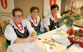 Kobiety w strojach stylizowanych na ludowe siedzą za białym stołem biesiadnym. Na stole widać dzbanek soku, p&oacute;łmiski z jedzeniem oraz sztućce. W tle banery z herbem oraz napisem &bdquo;Gmina Chork&oacute;wka&rdquo; i &bdquo;Koło Gospodyń Wiejskich&rdquo;.