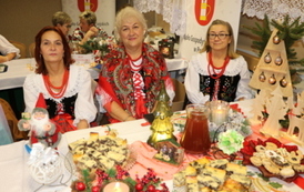 Kobiety w strojach stylizowanych na ludowe siedzą za białym stołem biesiadnym. Na stole widać dzbanek soku, p&oacute;łmiski z jedzeniem oraz sztućce. W tle banery z herbem oraz napisem &bdquo;Gmina Chork&oacute;wka&rdquo; i &bdquo;Koło Gospodyń Wiejskich&rdquo;.