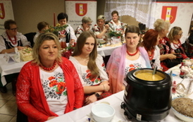 Kobiety w strojach stylizowanych na ludowe siedzą za białym stołem biesiadnym. Na stole widać dzbanek soku, p&oacute;łmiski z jedzeniem oraz sztućce. W tle banery z herbem oraz napisem &bdquo;Gmina Chork&oacute;wka&rdquo; i &bdquo;Koło Gospodyń Wiejskich&rdquo;.