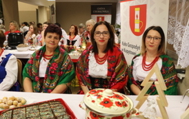 Kobiety w strojach stylizowanych na ludowe siedzą za białym stołem biesiadnym. Na stole widać dzbanek soku, p&oacute;łmiski z jedzeniem oraz sztućce. W tle banery z herbem oraz napisem &bdquo;Gmina Chork&oacute;wka&rdquo; i &bdquo;Koło Gospodyń Wiejskich&rdquo;.