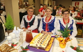 Kobiety w strojach stylizowanych na ludowe siedzą za białym stołem biesiadnym. Na stole widać dzbanek soku, p&oacute;łmiski z jedzeniem oraz sztućce. W tle banery z herbem oraz napisem &bdquo;Gmina Chork&oacute;wka&rdquo; i &bdquo;Koło Gospodyń Wiejskich&rdquo;.