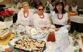 Kobiety w białych bluzkach siedzą za stołem na kt&oacute;rym stoją przygotowane potrawy. 