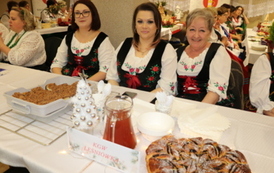 Kobiety w strojach stylizowanych na ludowe siedzą za białym stołem biesiadnym. Na stole widać dzbanek soku, p&oacute;łmiski z jedzeniem oraz sztućce. W tle banery z herbem oraz napisem &bdquo;Gmina Chork&oacute;wka&rdquo; i &bdquo;Koło Gospodyń Wiejskich&rdquo;.