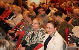 Rząd kobiet siedzących na widowni. Prawie każda z nich trzyma w dłoni pojedynczego tulipana owiniętego w folię. Kobiety są ubrane w kolorowe, eleganckie stroje i z uwagą śledzą program artystyczny.