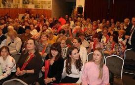 Widok z g&oacute;ry na wypełnioną po brzegi salę widowiskową. Publiczność, składająca się gł&oacute;wnie z kobiet, siedzi w rzędach na szarych krzesłach, uważnie obserwując scenę. Wnętrze ma drewniane panele na ścianach i parkietową podłogę.