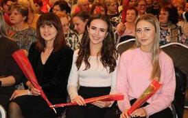 Rząd kobiet siedzących na widowni. Prawie każda z nich trzyma w dłoni pojedynczego tulipana owiniętego w folię. Kobiety są ubrane w kolorowe, eleganckie stroje i z uwagą śledzą program artystyczny.