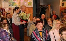 Widok z g&oacute;ry na wypełnioną po brzegi salę widowiskową. Publiczność, składająca się gł&oacute;wnie z kobiet, siedzi w rzędach na szarych krzesłach, uważnie obserwując scenę. Wnętrze ma drewniane panele na ścianach i parkietową podłogę.. Mężczyźni rozdają im kwiat