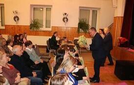 Widok z g&oacute;ry na wypełnioną po brzegi salę widowiskową. Publiczność, składająca się gł&oacute;wnie z kobiet, siedzi w rzędach na szarych krzesłach, uważnie obserwując scenę. Wnętrze ma drewniane panele na ścianach i parkietową podłogę.. Mężczyźni rozdają im kwiat