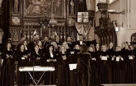 Szerokie ujęcie wnętrza zabytkowego kościoła w Korczynie, oświetlonego ciepłym światłem. Na stopniach przed monumentalnym, rzeźbionym, ciemnym ołtarzem stoi Ch&oacute;r Legato. Ch&oacute;rzyści (kobiety w długich czarnych sukniach, mężczyźni w garniturach) są ustawieni