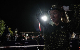 Kobieta w sportowej kurtce biegowej poprawia czoł&oacute;wkę na głowie w nocy, tuż przed startem biegu Ultra Trail Australia. W tle widać rozświetlone reflektorami miasteczko biegowe z kibicami.