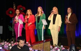 Występ wokalny zespołu kobiecego na scenie. Pięć kobiet ubranych w eleganckie, jednokolorowe garnitury w wyrazistych barwach (fuksja, czerwień, zieleń, ż&oacute;łć i brąz) śpiewa do mikrofon&oacute;w. W tle widoczne są dekoracje z motywem mak&oacute;w.
