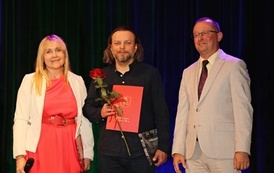 Uroczysty moment wręczenia wyr&oacute;żnienia. Mężczyzna trzyma a czerwoną r&oacute;żę oraz pamiątkowy dyplom stoi na scenie obok elegancko ubranego mężczyzny (przedstawiciela władz), kt&oacute;ry uśmiecha się w stronę publiczności.