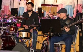 Muzycy akompaniujący artystom podczas koncertu. Na pierwszym planie widać perkusistę oraz basistę grających na swoich instrumentach. W tle, na podwyższeniu, widoczni są członkowie ch&oacute;ru oraz kwiatowe dekoracje sceny.