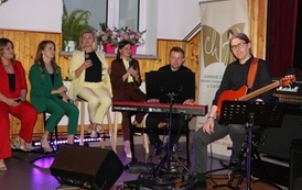 Muzycy akompaniujący artystom podczas koncertu. Na pierwszym planie widać perkusistę oraz basistę grających na swoich instrumentach. W tle, na podwyższeniu, widoczni są członkowie ch&oacute;ru oraz kwiatowe dekoracje sceny.