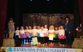 Zbliżenie na grupę dzieci stojących na scenie przed tłem przedstawiającym drzewa i światełka. Dzieci są ubrane w białe koszulki i sp&oacute;dniczki tiulowe w r&oacute;żnych jaskrawych kolorach (fioletowy, niebieski, ż&oacute;łty, r&oacute;żowy, zielony). Stoją przed mikrofonami, zapewne śpiewając.