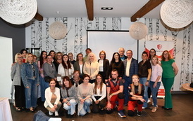 Zdjęcie grupowe wszystkich uczestnik&oacute;w szkolenia oraz instruktor&oacute;w, wykonane na koniec wydarzenia. Wszyscy stoją i klęczą, uśmiechnięci, patrząc w obiektyw aparatu. Tło stanowią charakterystyczna tapeta w brzozy, ekran projektora i baner reklamowy. Uczestnicy są w r&oacute;żnym wieku, ubrani w stroje codzienne. Zdjęcie upamiętnia zakończone szkolenie.