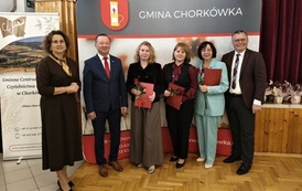 Zdjęcie pamiątkowe grupy os&oacute;b na tle ścianki Gminy Chork&oacute;wka. Trzy osoby w centrum trzymają czerwone teczki i żywe r&oacute;że. Grupie towarzyszą przedstawiciele samorządu oraz organizatorzy imprezy.