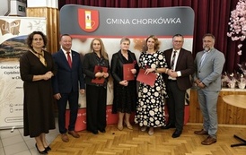 Pozujące do zdjęcia osiem os&oacute;b na tle ścianki Gminy Chork&oacute;wka. Trzy wyr&oacute;żnione kobiety w eleganckich kreacjach trzymają w rękach czerwone teczki pamiątkowe i kwiaty. W tle po prawej stronie widoczne jest dekoracyjne r&oacute;żowe drzewko.