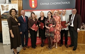 Osiem os&oacute;b pozuje na tle ścianki wystawienniczej Gminy Chork&oacute;wka. Wyr&oacute;żnione osoby trzymają czerwone teczki z nagrodami i czerwoną r&oacute;żę. W grupie znajdują się zar&oacute;wno kobiety, jak i mężczyzna odbierający wyr&oacute;żnienie.