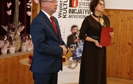 Mężczyzna w garniturze i czerwonym krawacie przemawia na pierwszym planie uroczystości. Obok niego stoi kobieta w ciemnej sukience, trzymająca czerwoną teczkę z dokumentami. W tle widoczne jest drewniane wykończenie sali, ozdobne drzewko oraz ścianka z napisem &bdquo;Wspieramy kulturę&rdquo;.
