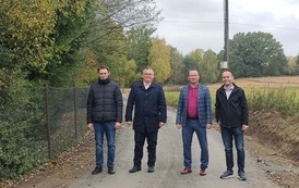 Czterech mężczyzn stoi w rzędzie na utwardzonej drodze gruntowej, patrząc w stronę aparatu. Są ubrani w odzież wierzchnią: od lewej czarna kurtka puchowa, bordowy sweter pod marynarką, czarny płaszcz, czerwono-czarna kurtka sportowa. Tło jest identyczne jak na poprzednim zdjęciu &ndash; wiejska droga prowadząca do oddalonej zabudowy z kościołem na wzg&oacute;rzu, otoczona polami. Niebo jest całkowicie zachmurzone.