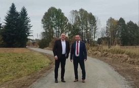 Dwaj mężczyźni stoją obok siebie na środku wąskiej, utwardzonej drogi żwirowej, prowadzącej w głąb kadru. Mężczyzna po lewej ma na sobie czarny garnitur, białą koszulę z rozpiętym kołnierzykiem i czarne buty. Mężczyzna po prawej ma na sobie ciemnoniebieski garnitur, białą koszulę i czerwony krawat oraz brązowe buty. Obaj patrzą w stronę aparatu.  Droga otoczona jest niską, wysuszoną roślinnością i pojedynczymi krzewami. W tle widać grupę liściastych i iglastych drzew, kt&oacute;rych liście mają jesienne, złoto-brązowe barwy. Niebo jest całkowicie zachmurzone i szare, tworząc płaskie oświetlenie. Zdjęcie zostało wykonane z perspektywy pieszego, patrzącego wzdłuż drogi.