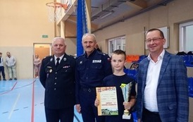 Zdjęcia przedstawiają uczestnik&oacute;w w strojach sportowych i strażackich podczas rywalizacji na hali sportowej, m.in. dynamiczne konkurencje z wężami strażackimi, uroczystą zbi&oacute;rkę wszystkich drużyn, a także momenty wręczania puchar&oacute;w, dyplom&oacute;w oraz nagr&oacute;d indywidualnych przez zaproszonych gości i przedstawicieli straży pożarnej.