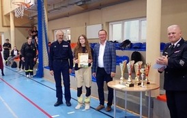 Zdjęcia przedstawiają uczestnik&oacute;w w strojach sportowych i strażackich podczas rywalizacji na hali sportowej, m.in. dynamiczne konkurencje z wężami strażackimi, uroczystą zbi&oacute;rkę wszystkich drużyn, a także momenty wręczania puchar&oacute;w, dyplom&oacute;w oraz nagr&oacute;d indywidualnych przez zaproszonych gości i przedstawicieli straży pożarnej.