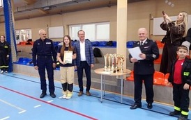 Zdjęcia przedstawiają uczestnik&oacute;w w strojach sportowych i strażackich podczas rywalizacji na hali sportowej, m.in. dynamiczne konkurencje z wężami strażackimi, uroczystą zbi&oacute;rkę wszystkich drużyn, a także momenty wręczania puchar&oacute;w, dyplom&oacute;w oraz nagr&oacute;d indywidualnych przez zaproszonych gości i przedstawicieli straży pożarnej.
