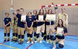 Zdjęcia przedstawiają uczestnik&oacute;w w strojach sportowych i strażackich podczas rywalizacji na hali sportowej, m.in. dynamiczne konkurencje z wężami strażackimi, uroczystą zbi&oacute;rkę wszystkich drużyn, a także momenty wręczania puchar&oacute;w, dyplom&oacute;w oraz nagr&oacute;d indywidualnych przez zaproszonych gości i przedstawicieli straży pożarnej.