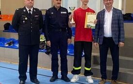 Zdjęcia przedstawiają uczestnik&oacute;w w strojach sportowych i strażackich podczas rywalizacji na hali sportowej, m.in. dynamiczne konkurencje z wężami strażackimi, uroczystą zbi&oacute;rkę wszystkich drużyn, a także momenty wręczania puchar&oacute;w, dyplom&oacute;w oraz nagr&oacute;d indywidualnych przez zaproszonych gości i przedstawicieli straży pożarnej.