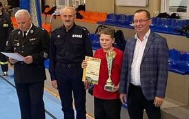 Zdjęcia przedstawiają uczestnik&oacute;w w strojach sportowych i strażackich podczas rywalizacji na hali sportowej, m.in. dynamiczne konkurencje z wężami strażackimi, uroczystą zbi&oacute;rkę wszystkich drużyn, a także momenty wręczania puchar&oacute;w, dyplom&oacute;w oraz nagr&oacute;d indywidualnych przez zaproszonych gości i przedstawicieli straży pożarnej.